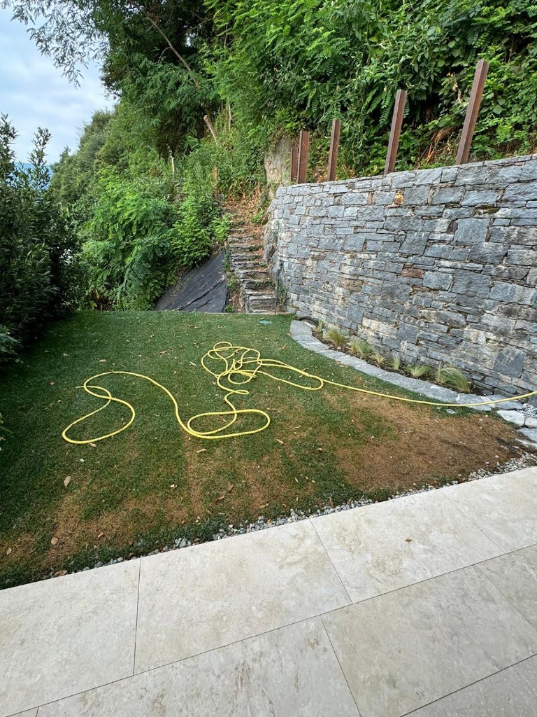 Rasen mit vergilbender Stelle und Gartenschlauch in einem Garten mit Steinmauer am Lago Maggiore